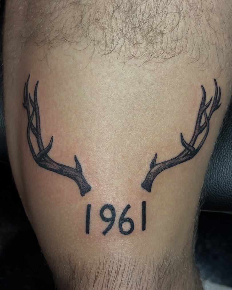 Solid black minimalist deer antlers tattooed symmetrically above the bold, clean numerals 1961 on a hairy leg or arm.