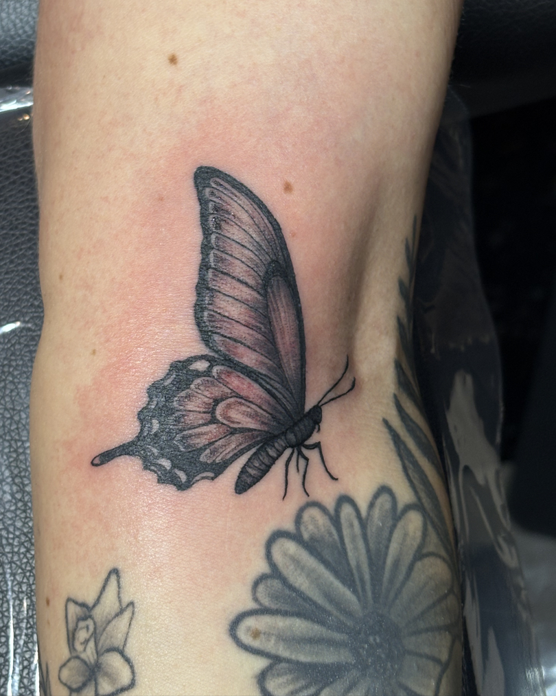 Detailed black and grey realistic butterfly tattoo with slightly pink-tinted wings, placed on the arm near other flower tattoos.