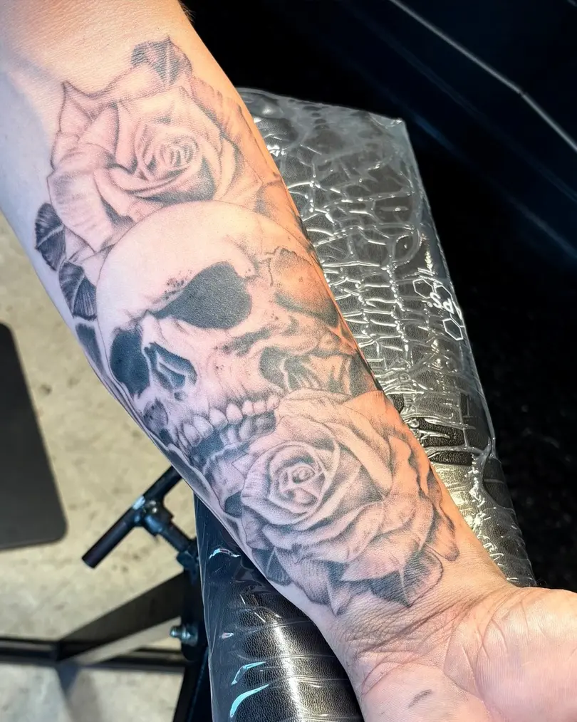 Detailed realistic black and grey tattoo of a human skull surrounded by large roses and foliage, running the length of the client's forearm.