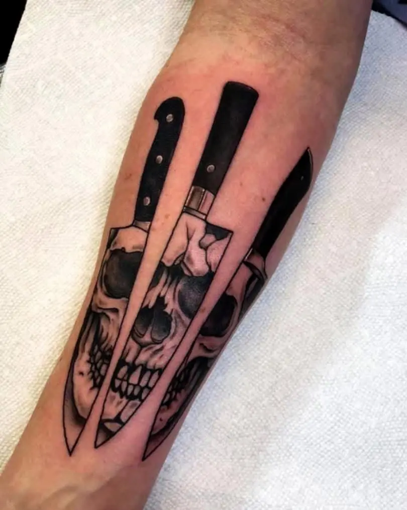 Three realistic black and grey kitchen knives tattooed on the forearm, with human skulls subtly reflected in the overlapping blades.