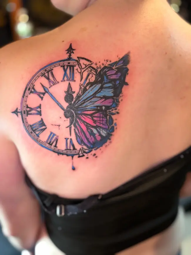 A back tattoo featuring half a Roman numeral clock and half a vibrant blue and pink butterfly wing. A semicolon symbol is at the center of the clock hands. The design includes artistic ink splatters and watercolor effects.