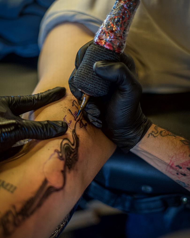 Close-up of a tattoo artist wearing black gloves holding a machine, outlining fine line script and flowing illustration on a client's inner forearm during a tattooing session.